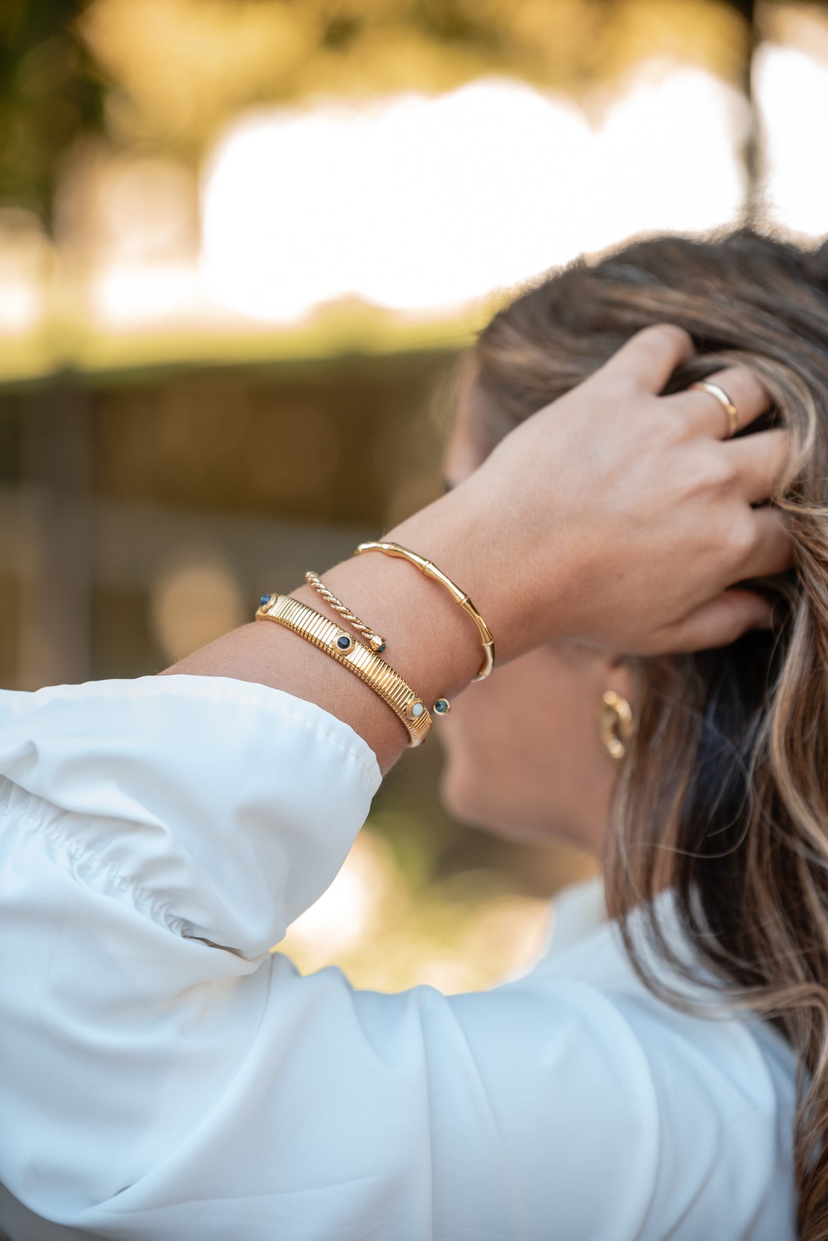 Pulso da Carmo com três pulseiras douradas; a pulseira dourada larga e canelada, com pedras azuis e brancas é a mais larga